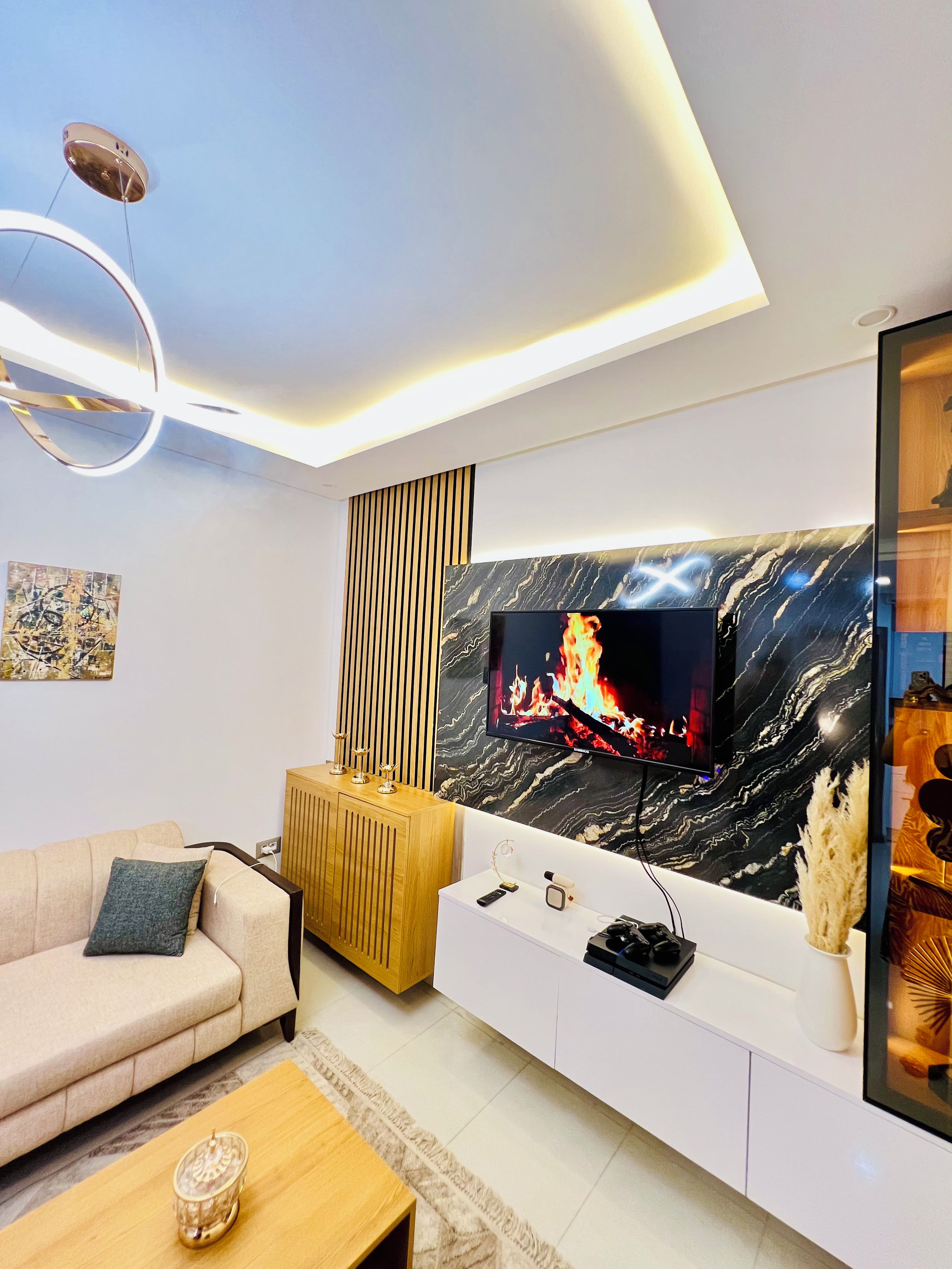 Ein modernes Wohnzimmer mit einem flachbildfernseher an einer Wand mit Marmormuster, einer Holzkommode, einem beigefarbenen Sofa und einem Couchtisch.