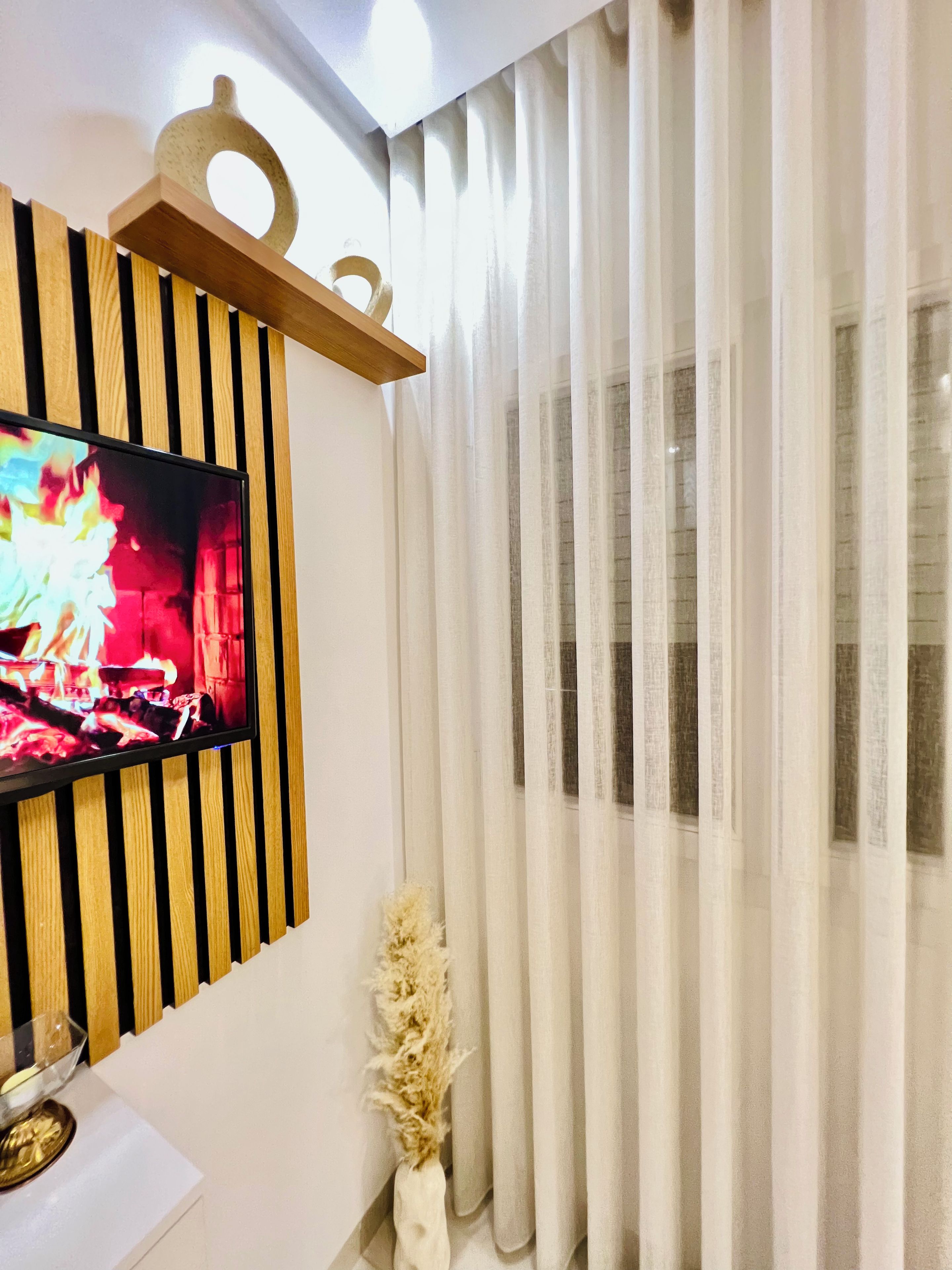 Una sala de estar moderna con un televisor de pantalla plana montado en un panel de madera, un jarrón blanco con plantas secas y cortinas blancas traslúcidas.