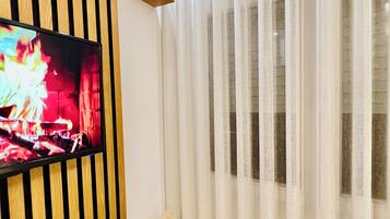 Una sala de estar moderna con un televisor de pantalla plana montado en un panel de madera, un jarrón blanco con plantas secas y cortinas blancas traslúcidas.