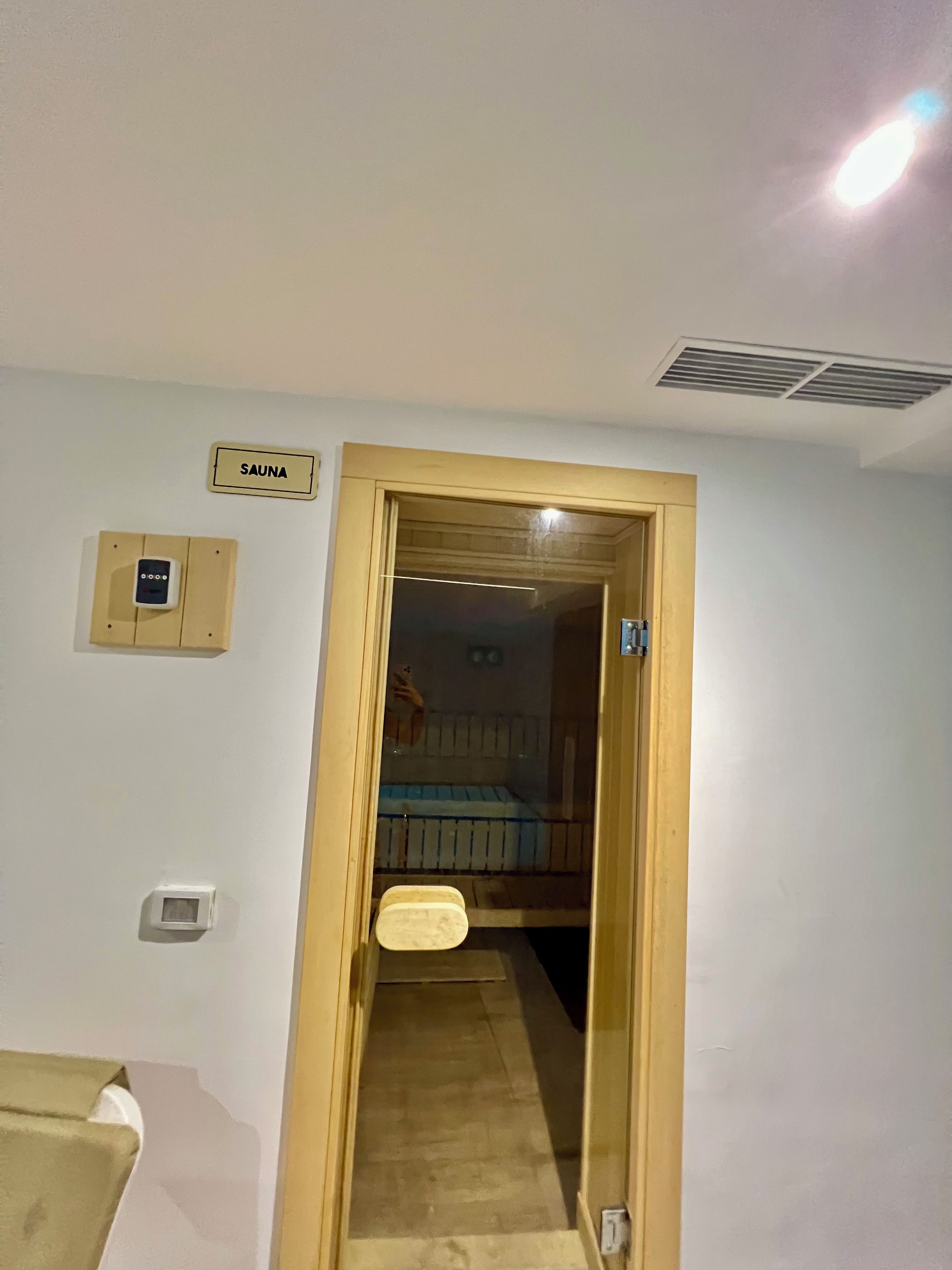 Una stanza con una porta in legno che dà su un'area sauna, un piccolo pannello di controllo e una zona doccia visibile.