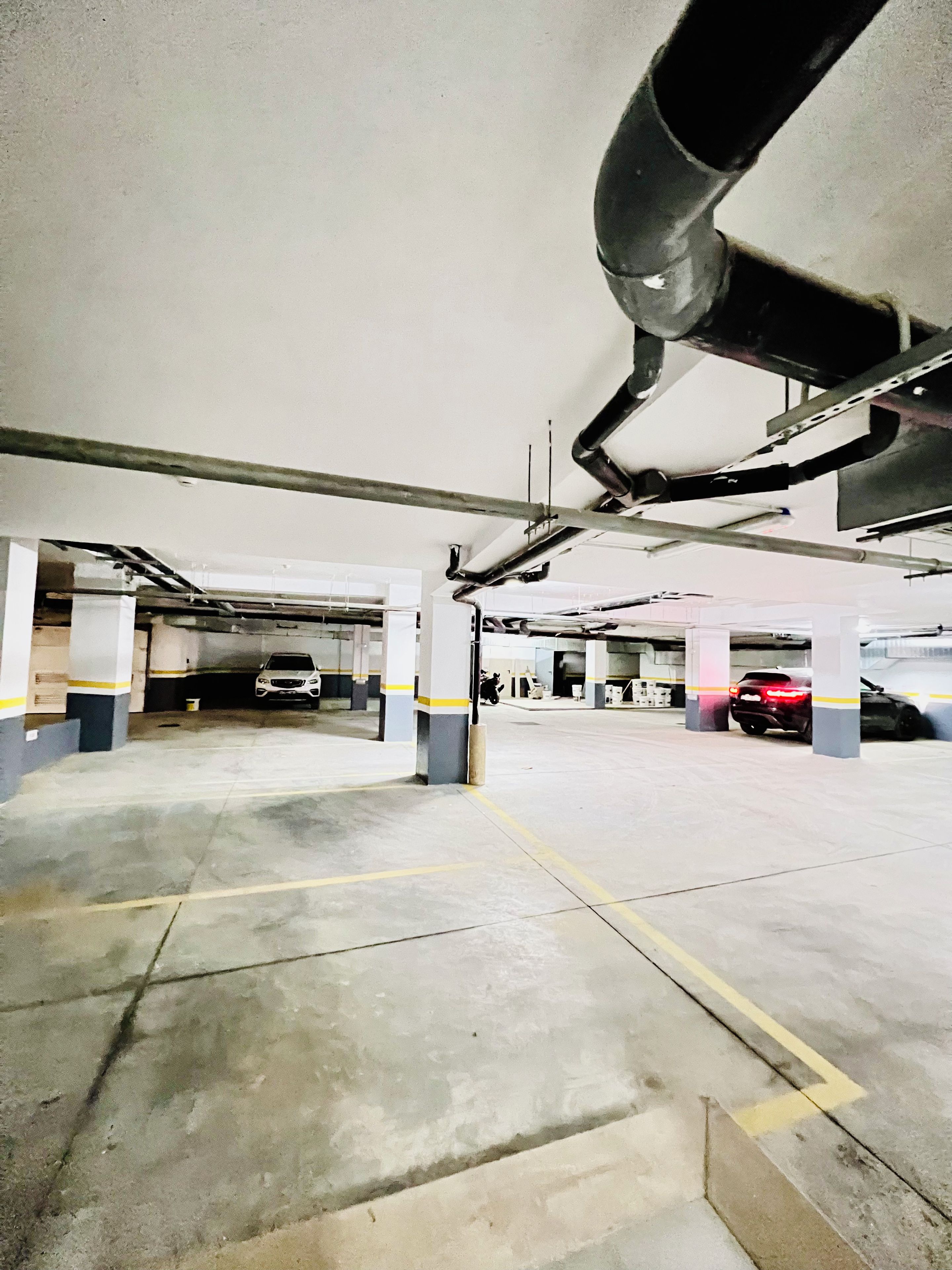 Een meerlaagse parkeergarage met betonnen vloeren en kolommen, met enkele geparkeerde voertuigen en zichtbare constructieelementen zoals leidingen en balken.
