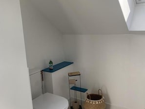 Een kleine, witte badkamer met een toilet, een plankje met een fles, een blauwe plank, een mand en een dakraam.