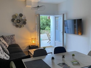 Ein modernes Wohnzimmer mit einer Couch, einem Esstisch mit Stühlen, einem an der Wand befestigten Fernseher und einer Tür zu einem Außenbereich.