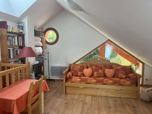A cozy attic bedroom with a sloped ceiling, a wooden sofa with patterned cushions, a small table with a red tablecloth, and a bookshelf filled with books. - Charming duplex apartment - 6 guests (Valloire)