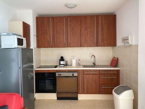 Cocina compacta con armarios de madera, microondas, refrigerador, lavavajillas y un basurero.