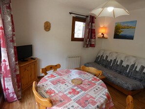 Ein gemütliches Wohnzimmer mit einem runden Tisch, Holzstühlen, einem Fernseher auf einer Holzkommode, einem Sofa mit Stadtblick-Motiv und einem Bild an der Wand.