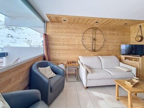 Ein modernes Wohnzimmer mit Holzboden, einem großen Fenster, einer Couch, einem Sessel, einem Fernseher und einem Holztisch.