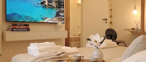 A hotel room with a bed, a TV displaying a scenic view, a bedside table with books, and a tray with cups and a vase.