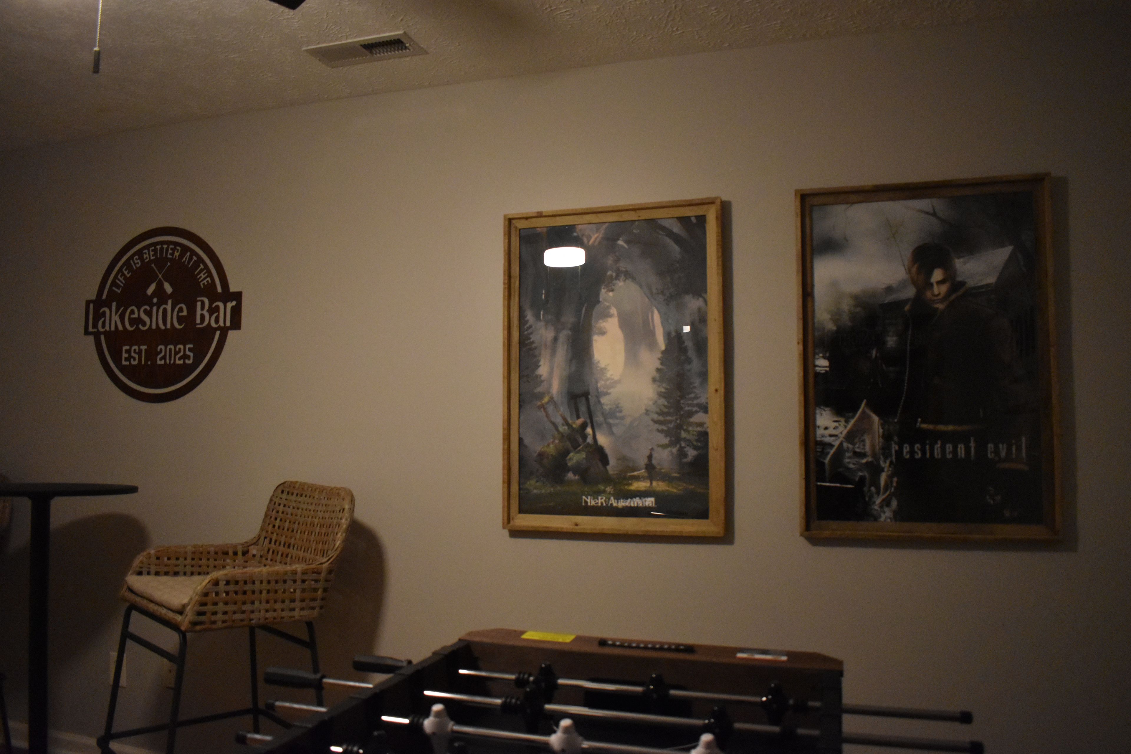 A bar area with a foosball table, a wicker chair, and two framed posters on the wall.