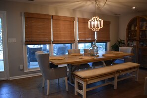 A dining area with a wooden table, chairs, and a bench. There are large windows with blinds, a chandelier, and a cabinet with decorative items.
