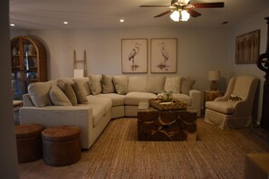 A living room with a sectional sofa, a coffee table, a side table, and a chair. There are two framed pictures on the wall, a ceiling fan, and a lamp on a side table.