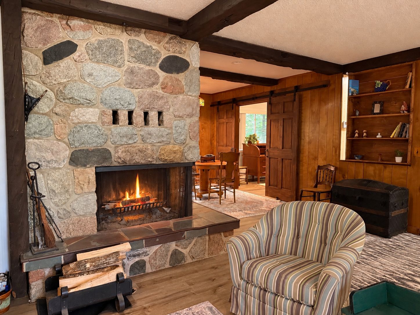 Una chimenea de piedra con fuego, un sillón a rayas, y estanterías de madera con libros y objetos decorativos.