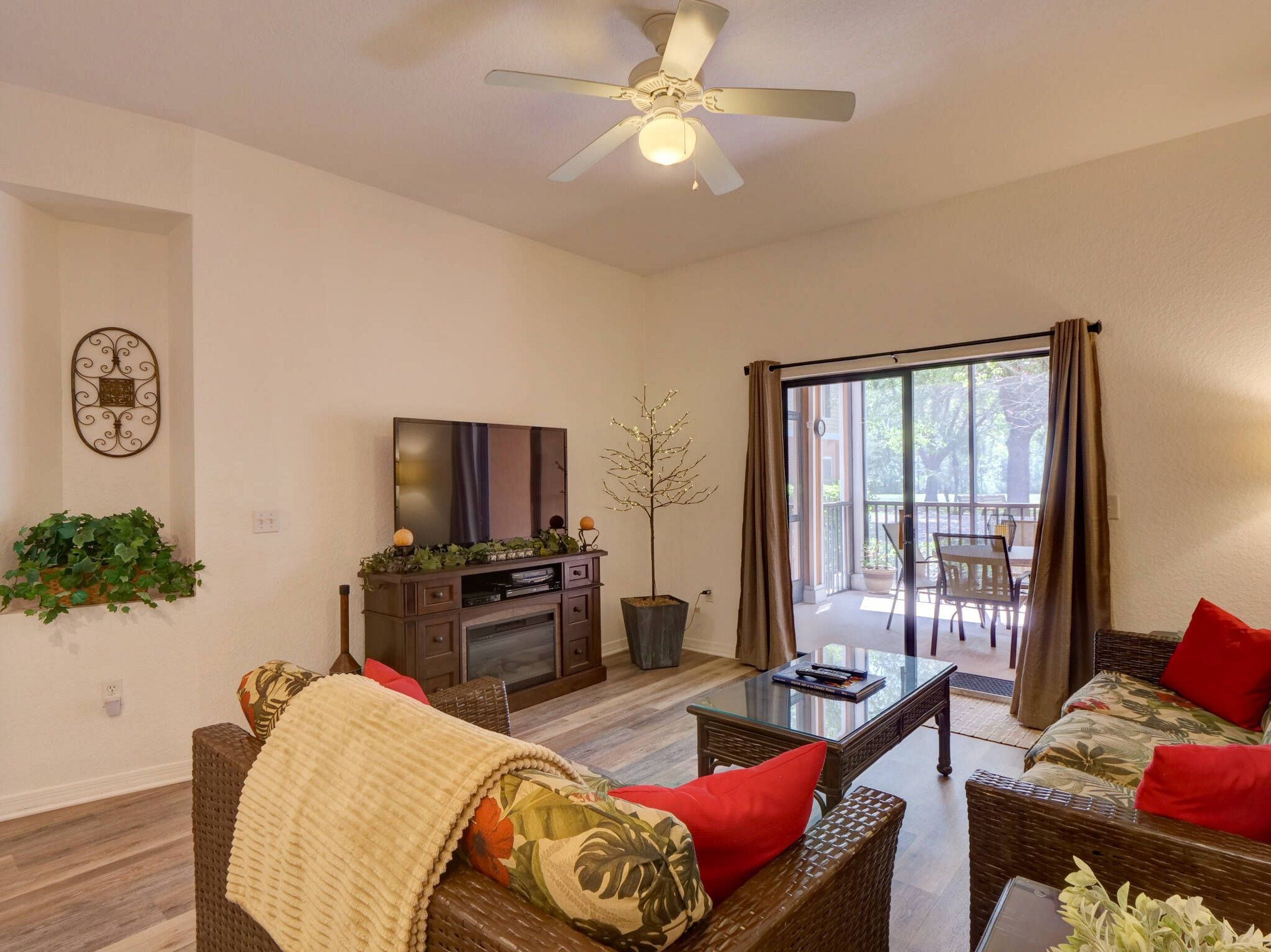 A living room with a ceiling fan, a flat-screen TV, a coffee table, wicker furniture, and a sliding glass door leading to an outdoor area.