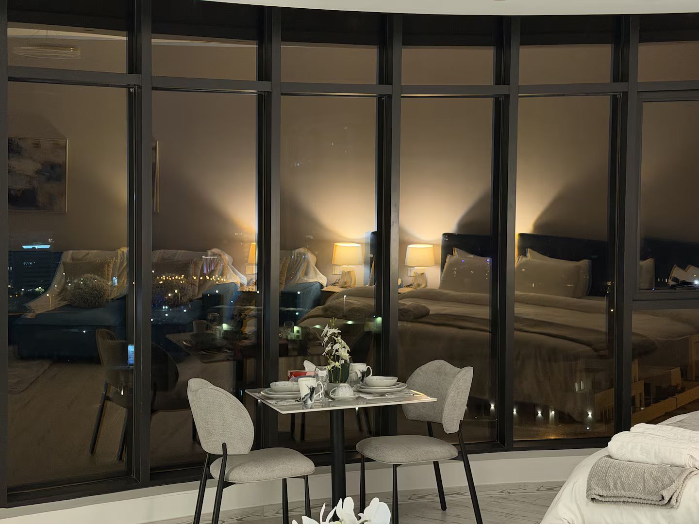 A modern hotel room with a large window, a dining area with a table and chairs, and a bed with a headboard.