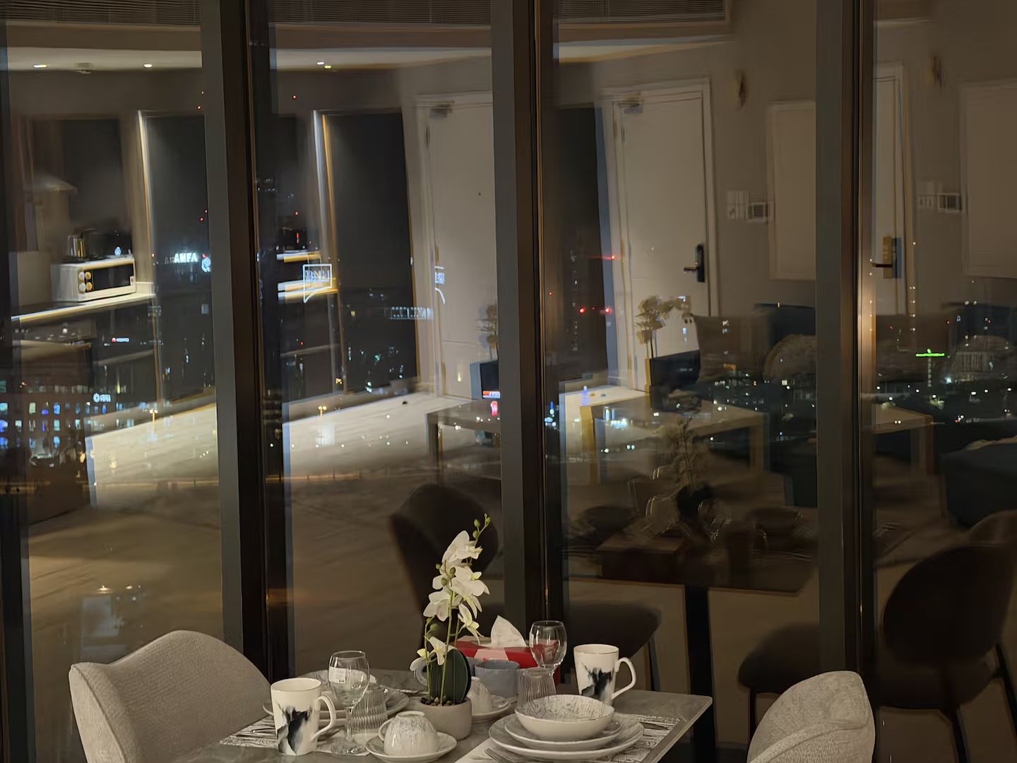 A dining area with a table set for two, a vase of white flowers, and a city view through large windows.