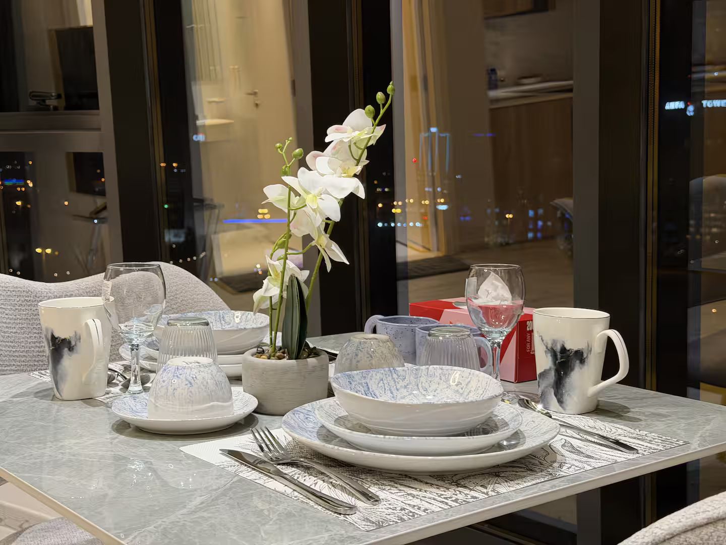 A table set for dining with plates, bowls, glasses, and cutlery, and a potted orchid in the center.