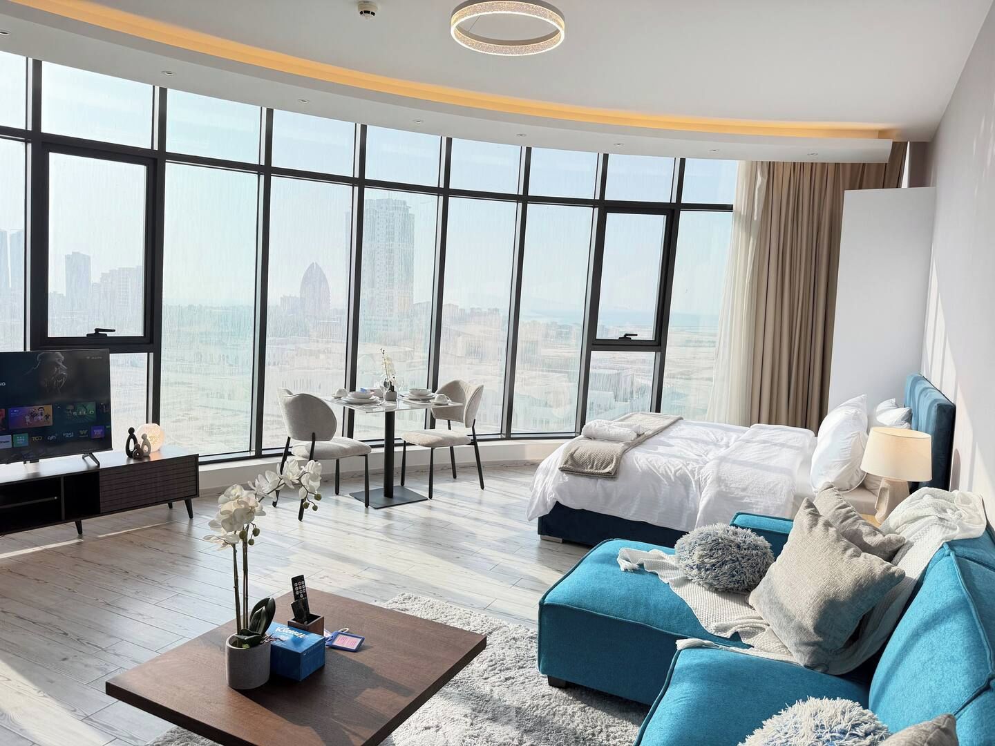 A modern hotel room with a large window, a bed, a seating area, and a television.