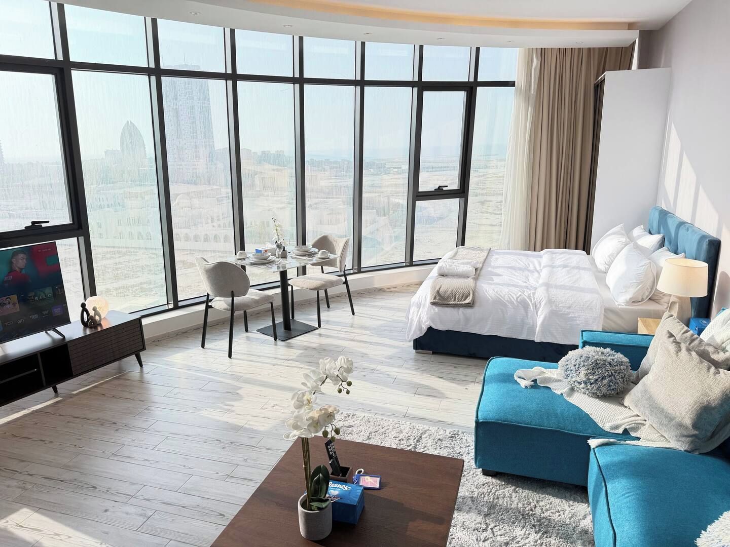 A modern hotel room with a large window, a bed, a sofa, a TV, and a dining area.