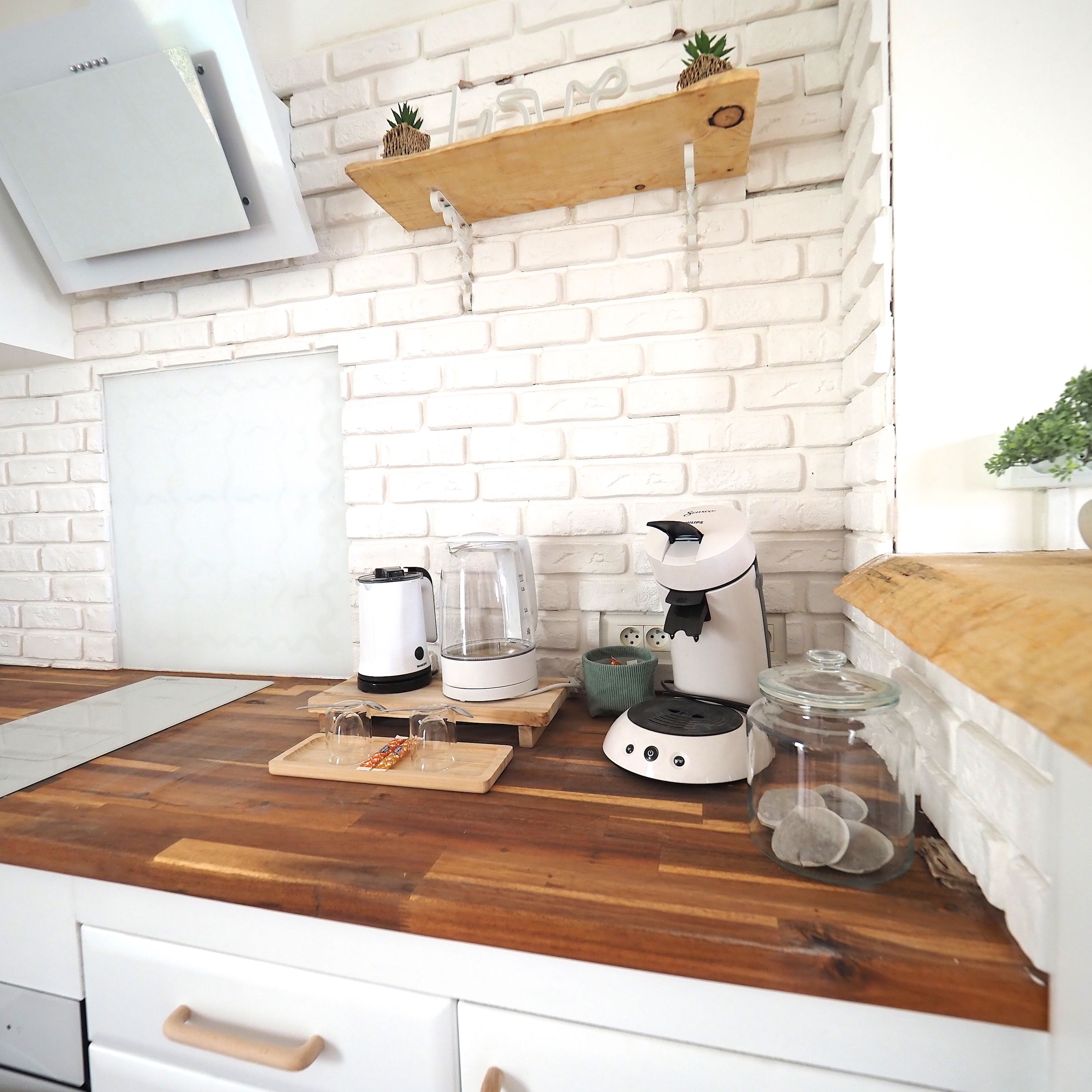 A kitchen with white brick walls, a wooden countertop, a white air conditioner, a coffee maker, a kettle, a glass container, and a shelf with a pineapple.