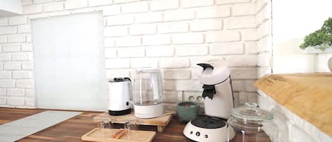 A kitchen with white brick walls, a wooden countertop, a white air conditioner, a coffee maker, a kettle, a glass container, and a shelf with a pineapple.