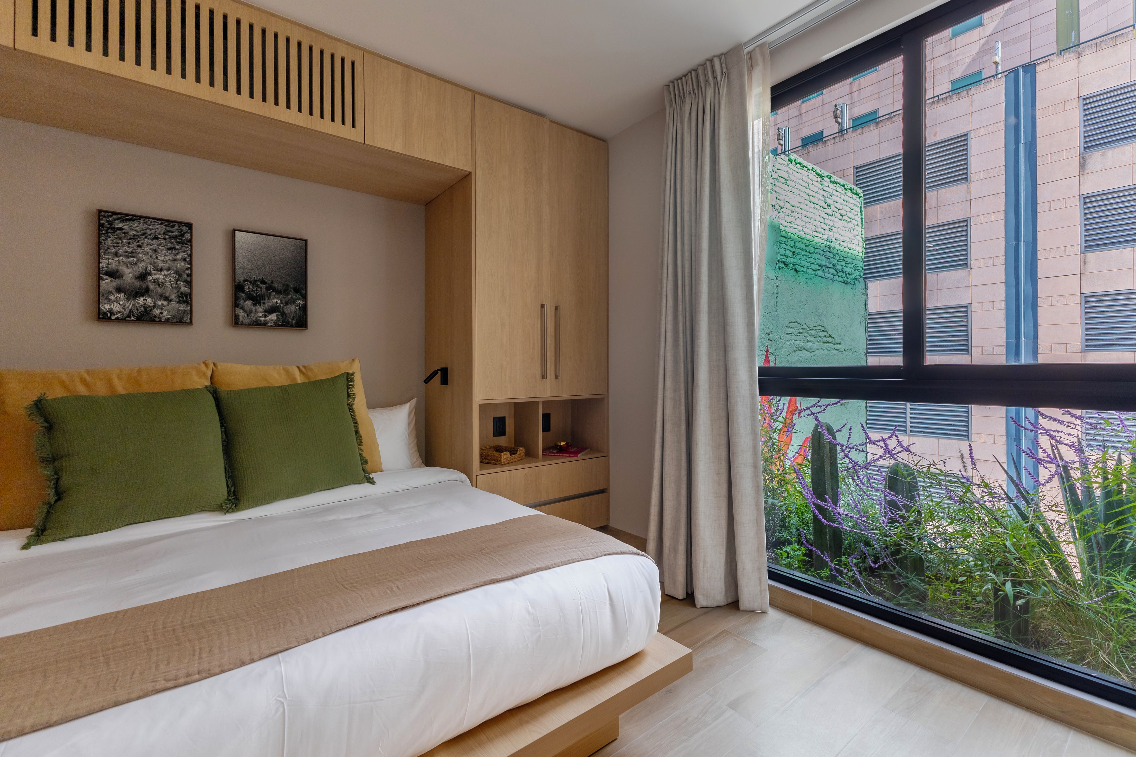 A bedroom with a large bed, wooden headboard, two framed pictures, a built-in wardrobe, and a window with a view of buildings and greenery.