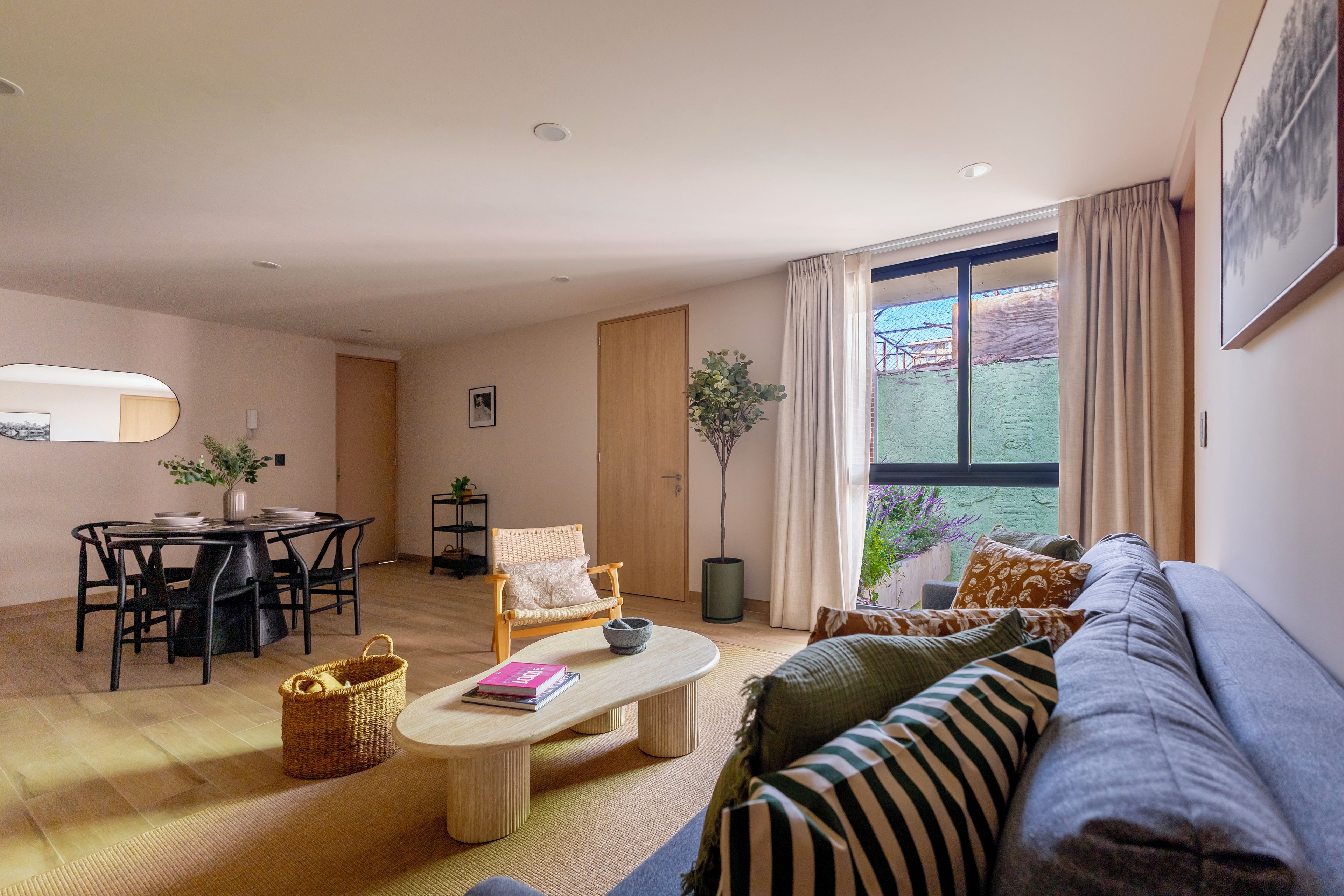A modern living room with a dining area, a large window with curtains, and a view of greenery outside.