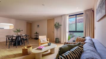 A modern living room with a dining area, a large window with curtains, and a view of greenery outside.