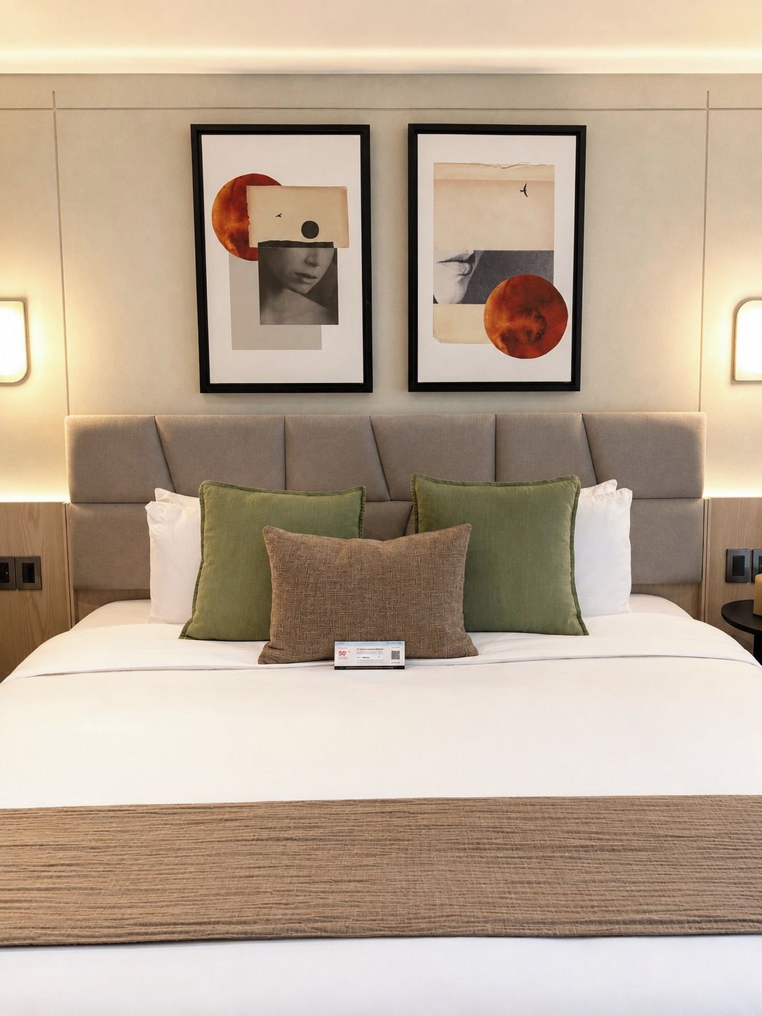 A neatly made bed with a pillow and a remote control, a headboard with two framed artworks, and bedside lamps.