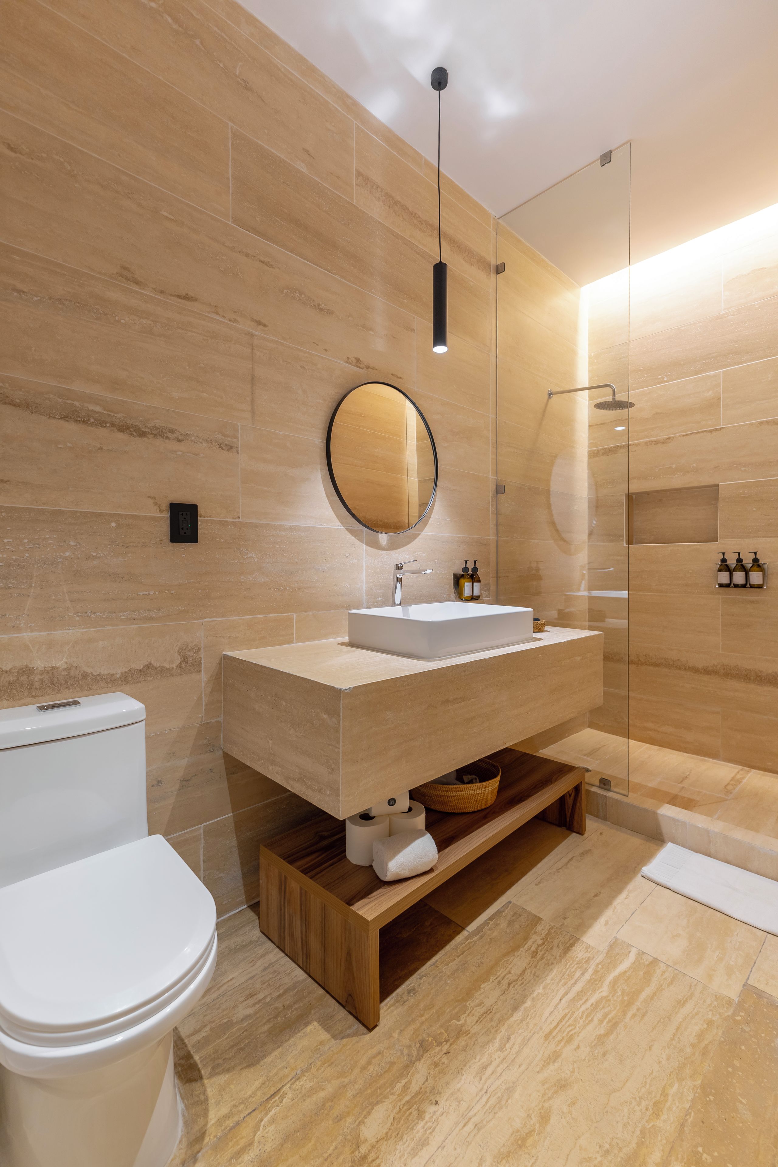 A modern bathroom with a wooden vanity, a round mirror, a toilet, and a glass-enclosed shower area.