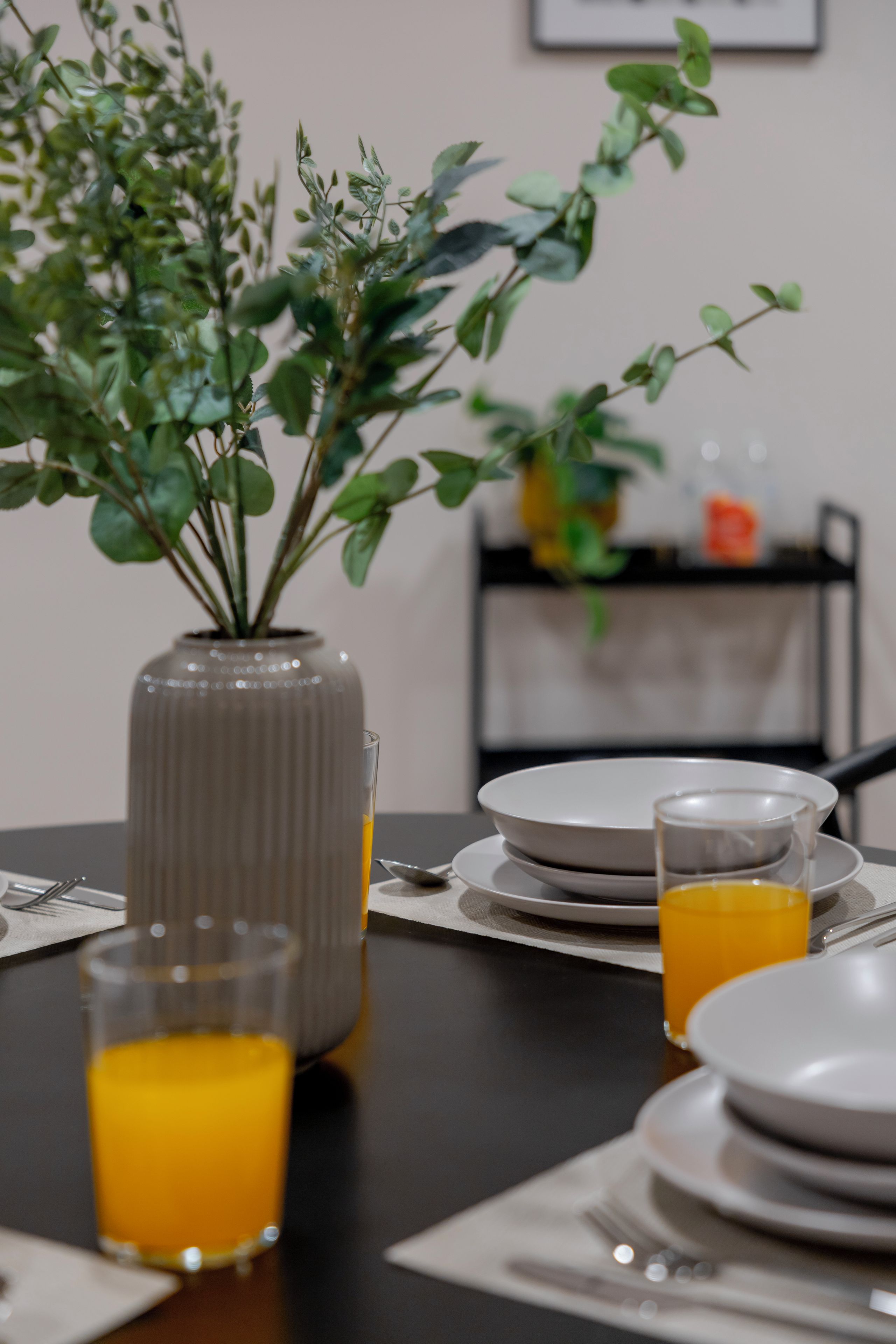 Una mesa de comedor con un florero de plantas verdes, dos vasos de jugo de naranja y platos blancos.