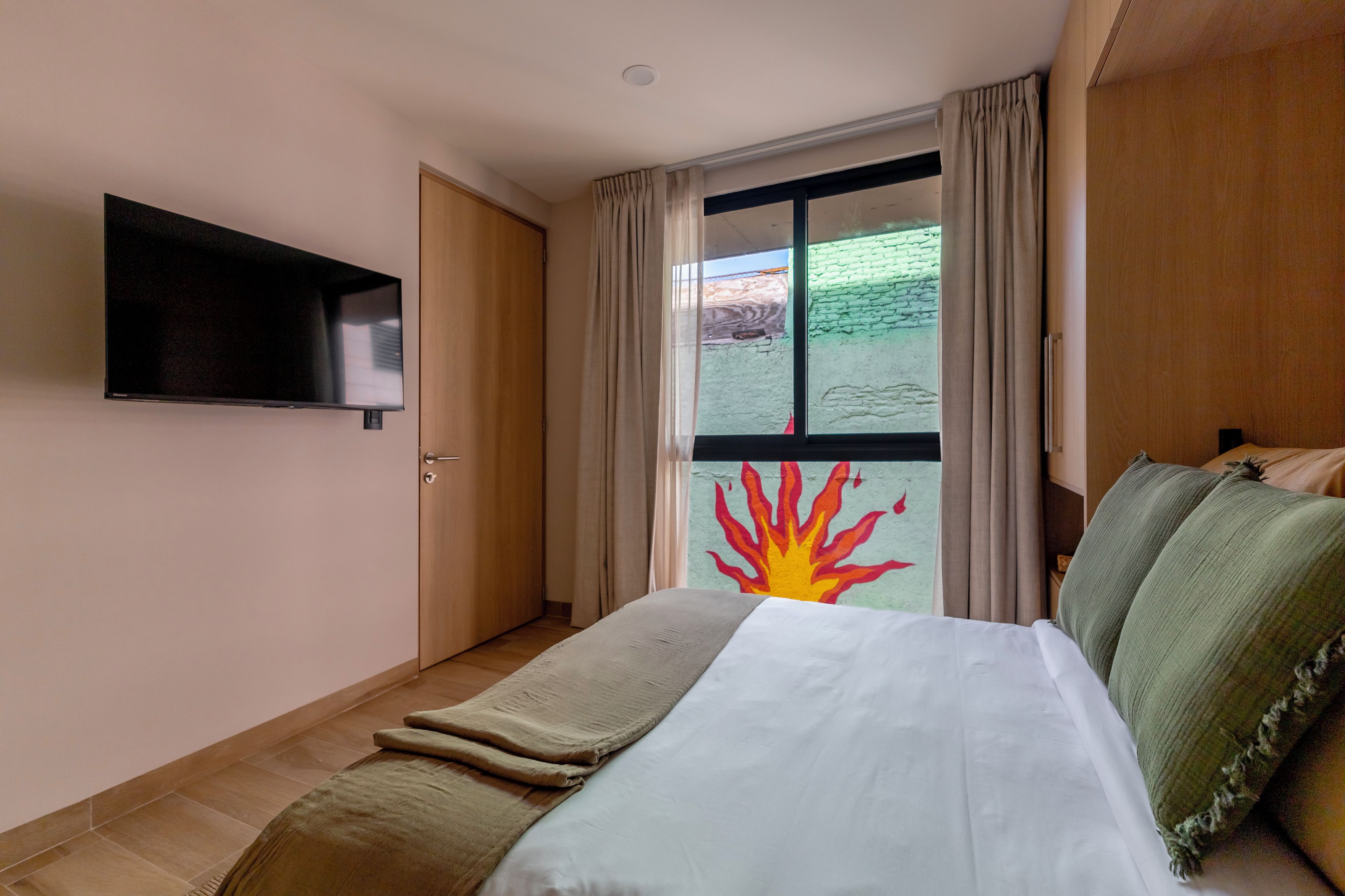Quarto de hotel com cama, TV de tela plana na parede, janela com cortinas e vista para um edifício com fachada verde.