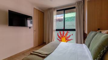 Quarto de hotel com cama, TV de tela plana na parede, janela com cortinas e vista para um edifício com fachada verde.