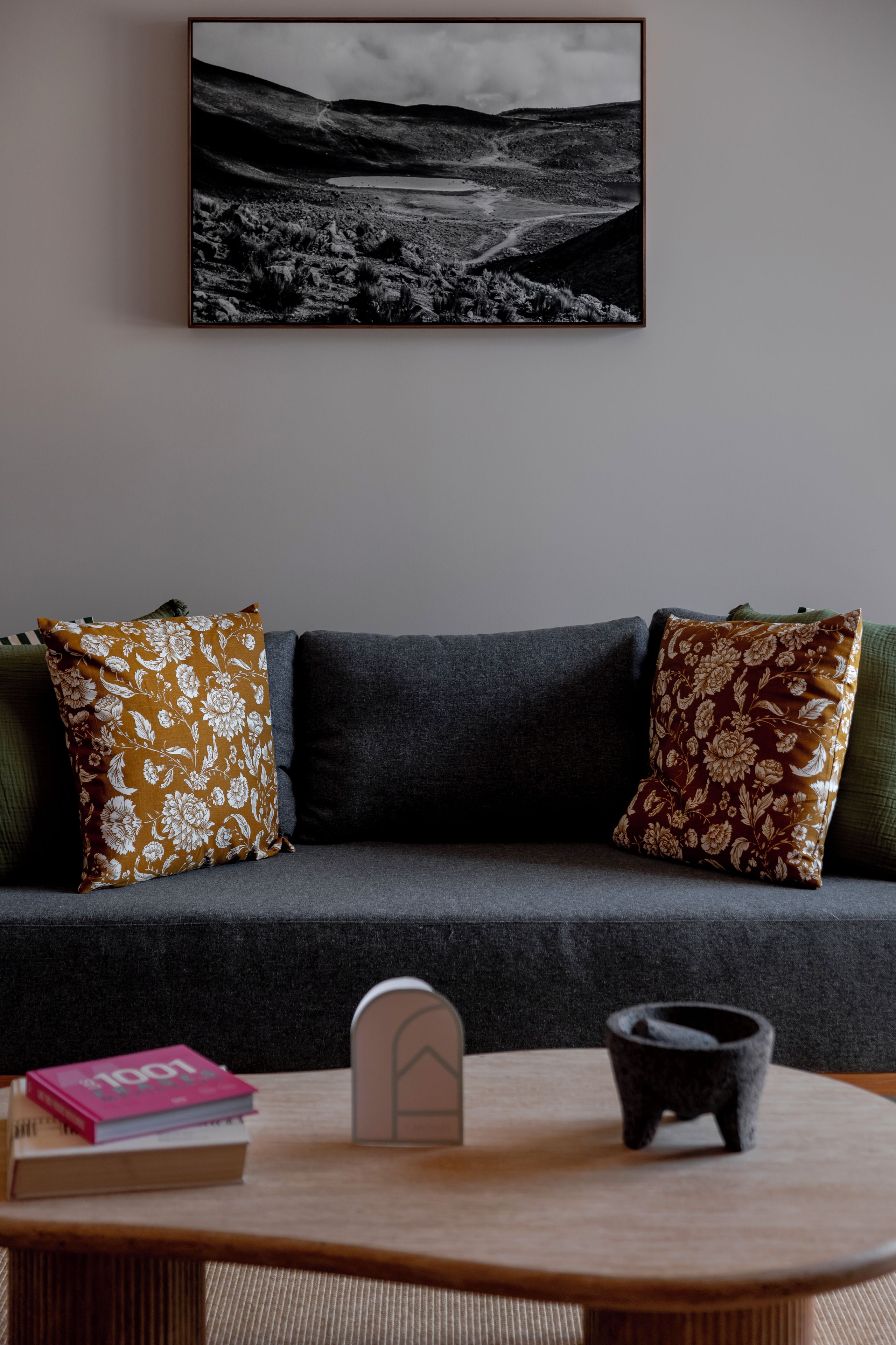 A modern living room with a grey sofa, two patterned throw pillows, a small wooden table with books and a decorative item, and a framed black and white photograph on the wall.