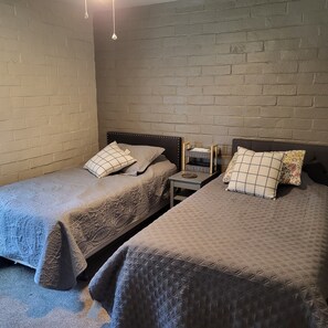 Ein Zimmer mit zwei Betten, jeweils mit Kopfteil und Kissen, einem kleinen Tisch mit Lampe und einer Ziegelwand.