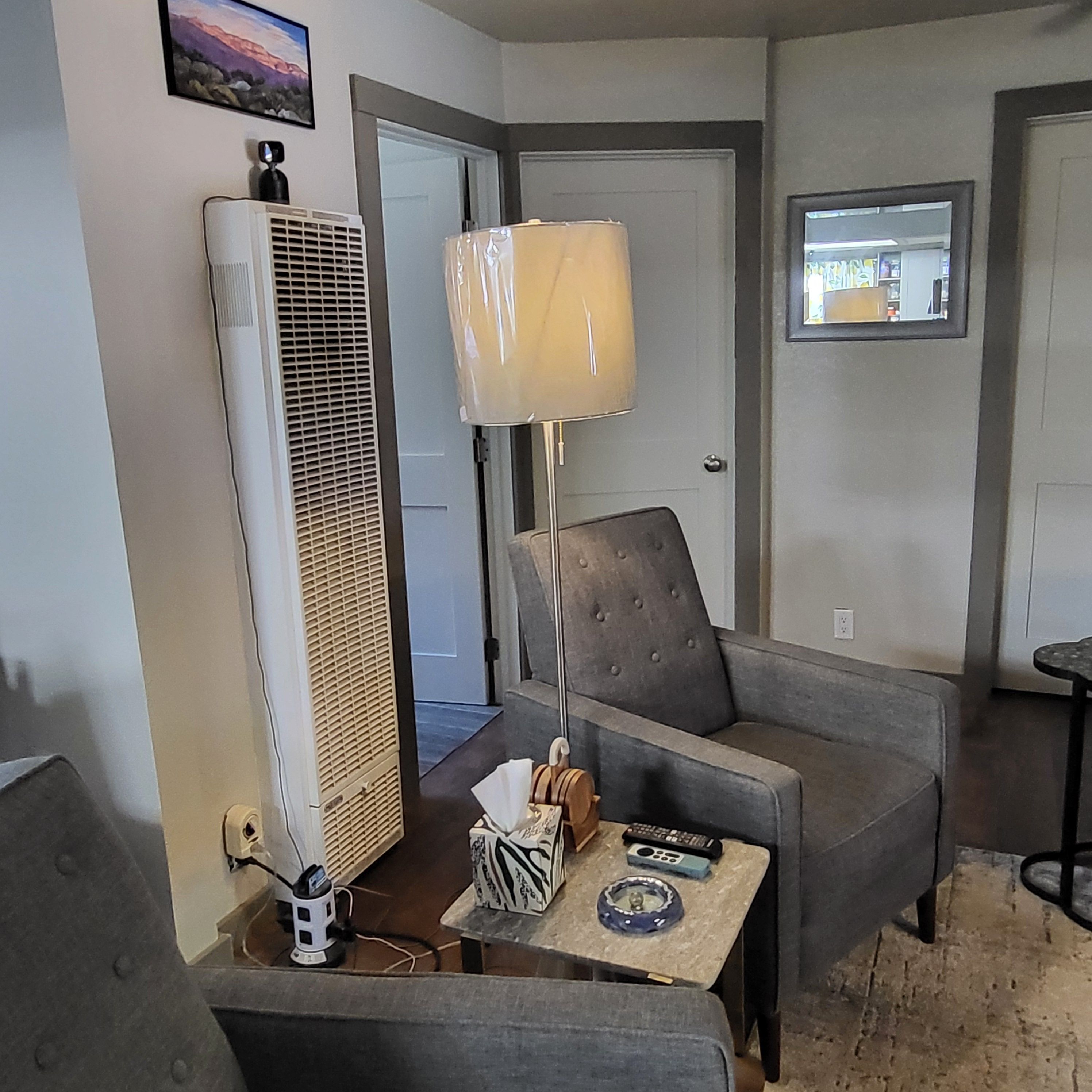 En stue med en grå lænestol, et lille bord med en lampe og en stor aircondition.