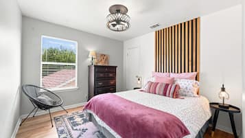 Quarto com cama, penteadeira, cadeira, janela com persianas e luminária de teto.