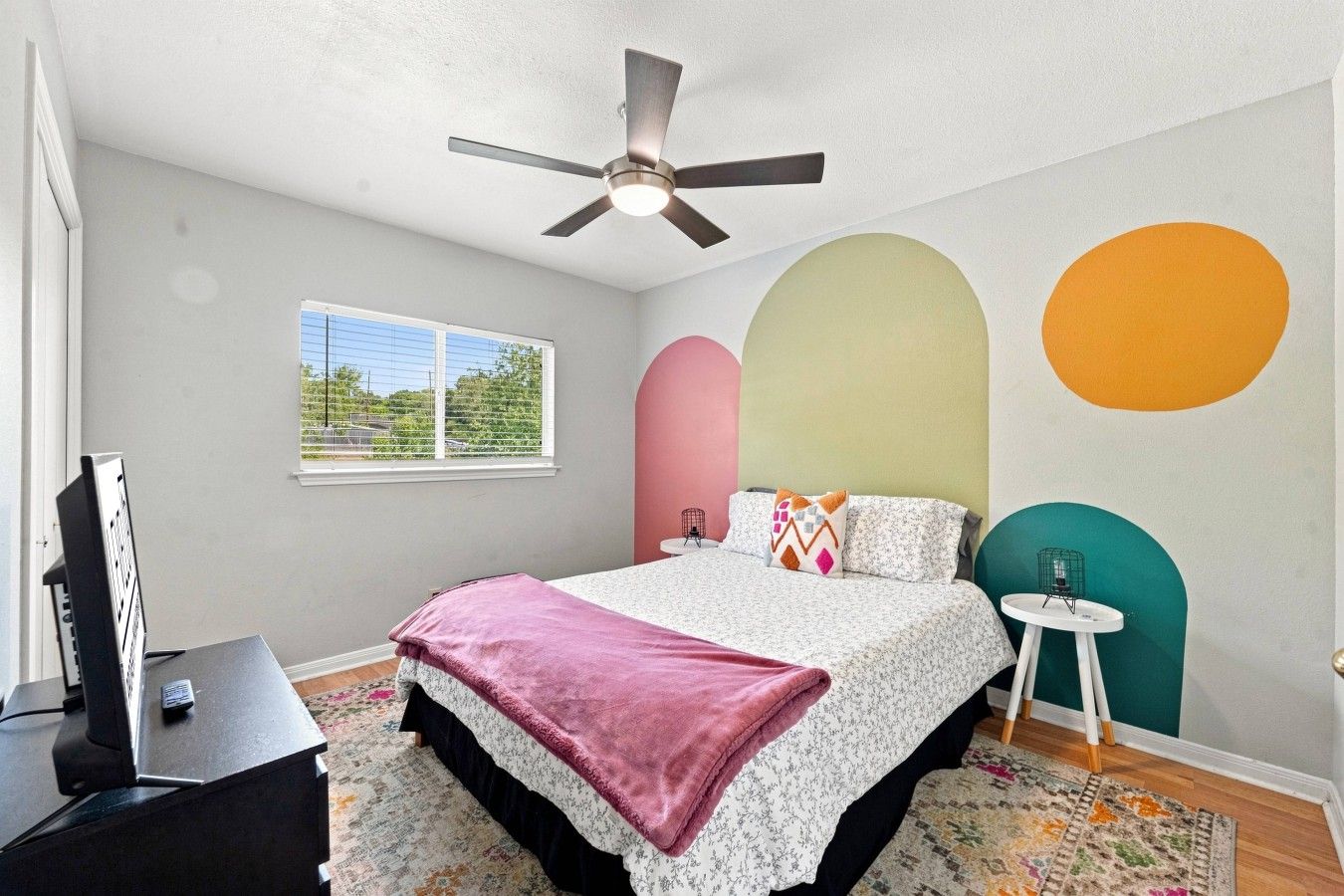A bedroom with a bed, a ceiling fan, a window, a desk, and a TV.