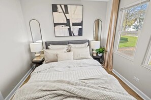 A bedroom with a bed, two bedside tables, a wall-mounted abstract painting, a window with curtains, and a view of the outdoors. - Relaxing getaway 3-bedroom 2-bathroom house in charming Crystal Lake (Crystal Lake)