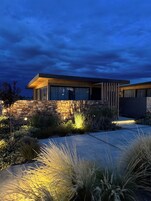 Una casa moderna con fachada de piedra, ventanas grandes y un jardín bien iluminado al anochecer.