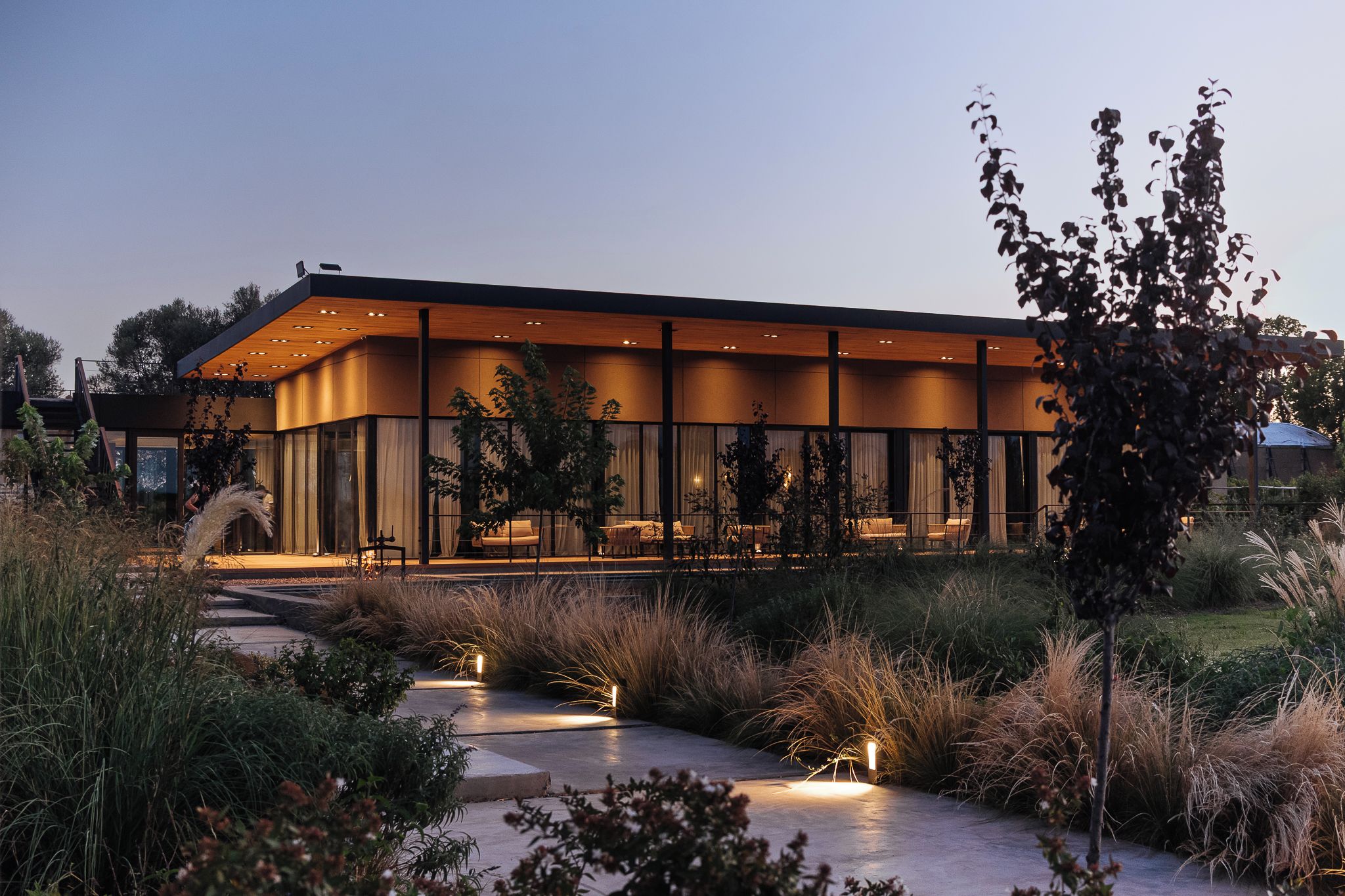 A modern building with a glass facade, a covered outdoor seating area, and a pathway leading to the entrance.