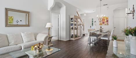 A modern living room with a glass coffee table, a white sofa, and a dining area with a chandelier.