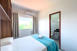 Une chambre d’hôtel comprenant un lit, une fenêtre avec des rideaux, un appareil de climatisation, une salle de bain avec un miroir et une porte.
