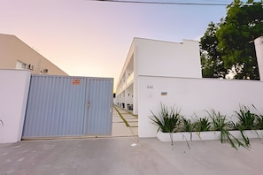 A gated entrance to a building with a white exterior, a blue shipping container, and a row of potted plants. - Apartamento 5 com 2 suítes 450m da praia (Santa Cruz Cabrália)