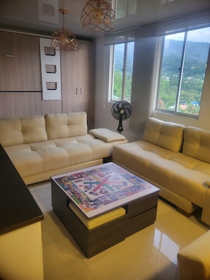 A modern living room with a sectional sofa, a coffee table with a board game, a standing fan, and large windows offering a view of the outdoors.