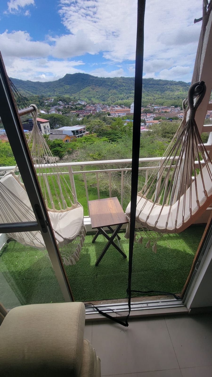 A room with a view of a town and mountains, a hammock chair, a small table, and a sofa.