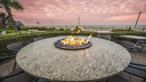 Ein runder Tisch im Freien mit einer Feuerstelle in der Mitte, umgeben von Stühlen, vor einer Kulisse aus Strand und Sonnenuntergangshimmel.