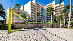 A beach volleyball court with a net in front of a multi-story hotel with balconies and palm trees.