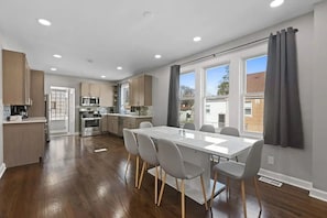 A modern kitchen with stainless steel appliances, a dining area with a white table and grey chairs, and large windows with curtains. - Stunning open-concept  3-bedroom 2-bathroom fully equipped home in Morton Grove (Morton Grove)