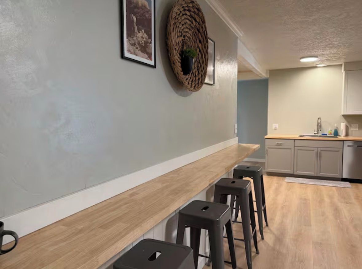 Eine Küche mit Holzauszug und Barhocker, einem hängenden gewebten Korb und einem gerahmten Bild an der Wand.