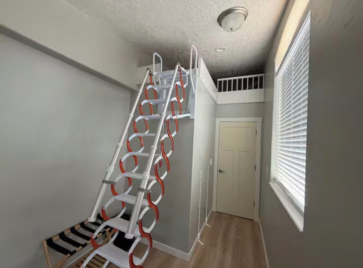 Eine Treppe mit Metallgeländer und orangefarbenen Akzenten, die zu einer Zwischenebene mit einer Tür und einem Fenster mit Jalousien führt.